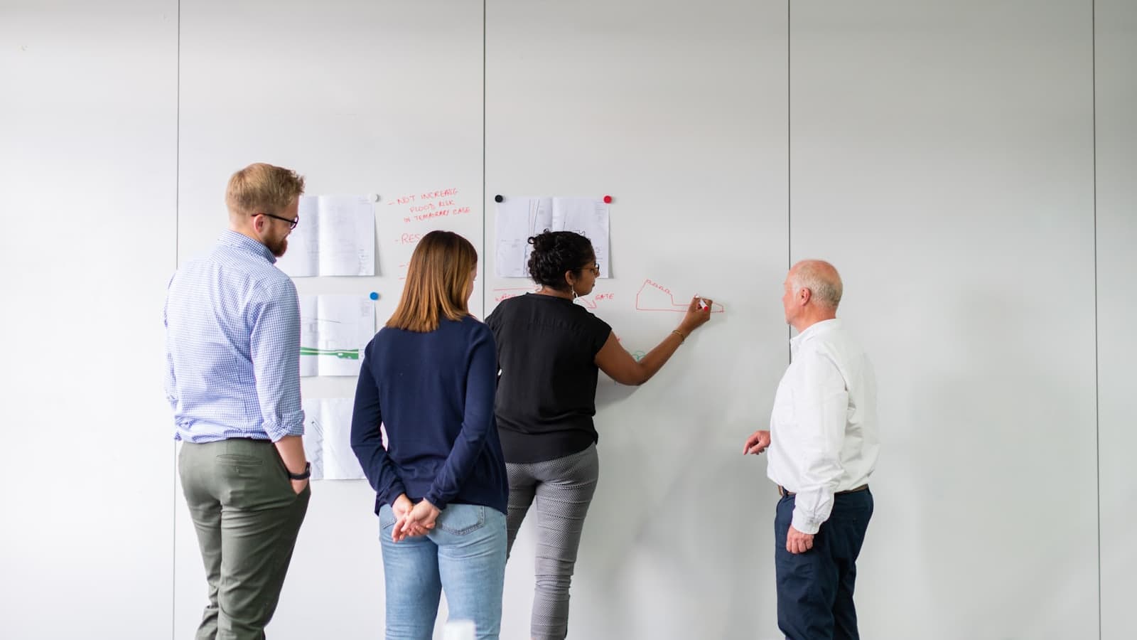 Four colleagues at a whiteboard wall hung with printed plans, one of them sketching in red marker.