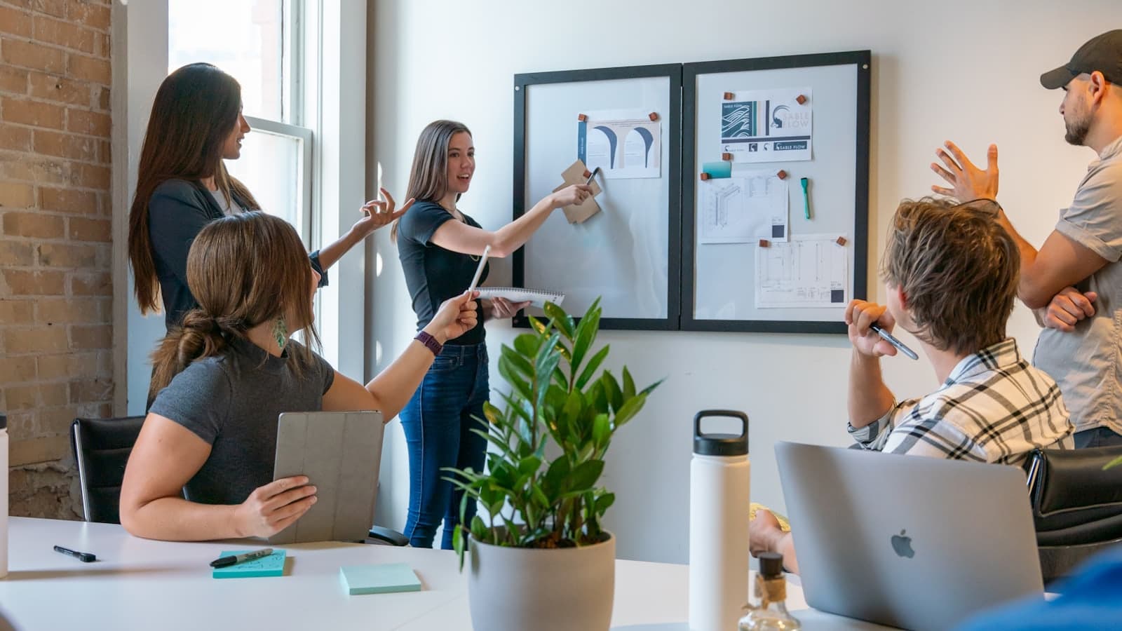 Colleagues around a table in a bright brick-walled studio, one of them pinning a document to a whiteboard while the others talk it through.