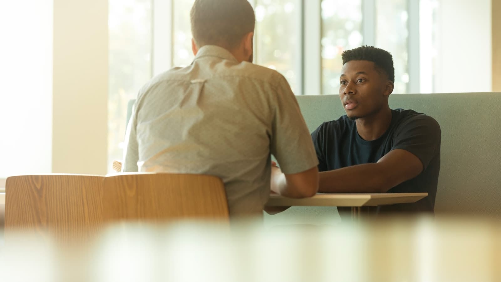 Two people talking across a table in a sunlit café booth, one listening while the other speaks.