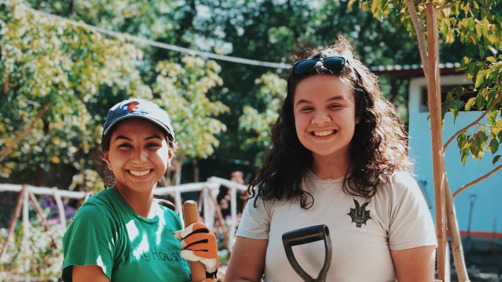 Two smiling volunteers holding garden tools beside a freshly planted sapling on a sunny community planting day.