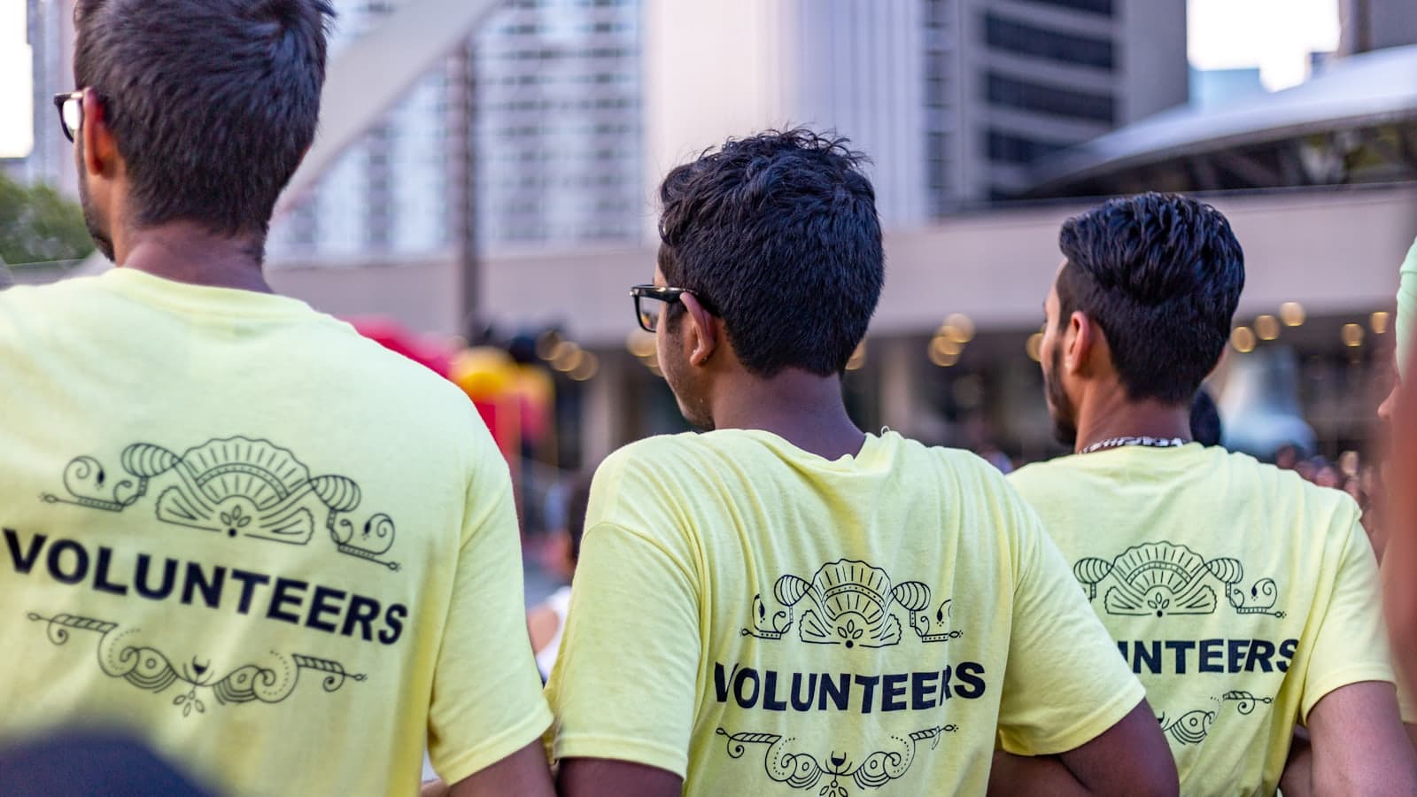 Three volunteers seen from behind at a city event, wearing pale yellow t-shirts printed with the word VOLUNTEERS.