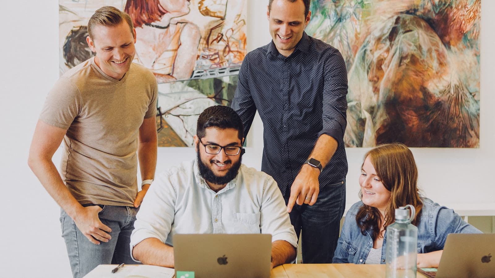 Four colleagues gathered around a laptop in a bright office, one of them pointing at the screen.