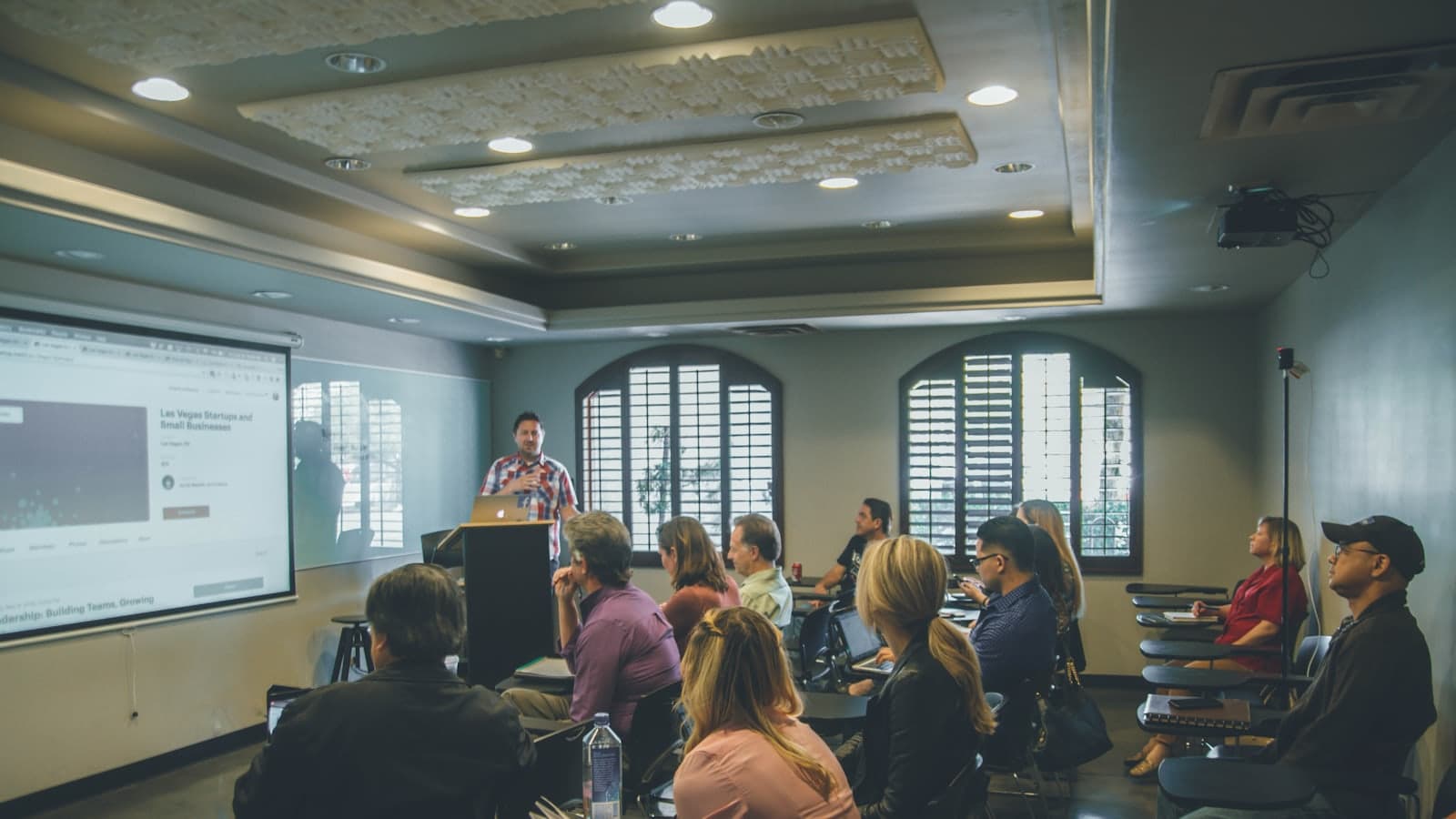 People seated around tables in a meeting room, listening to a facilitator presenting beside a projector screen.